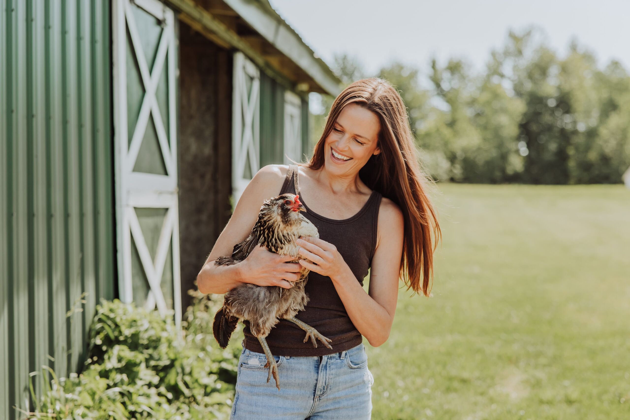 Kelli smiling while holding a chicken beside the green barn at her farm